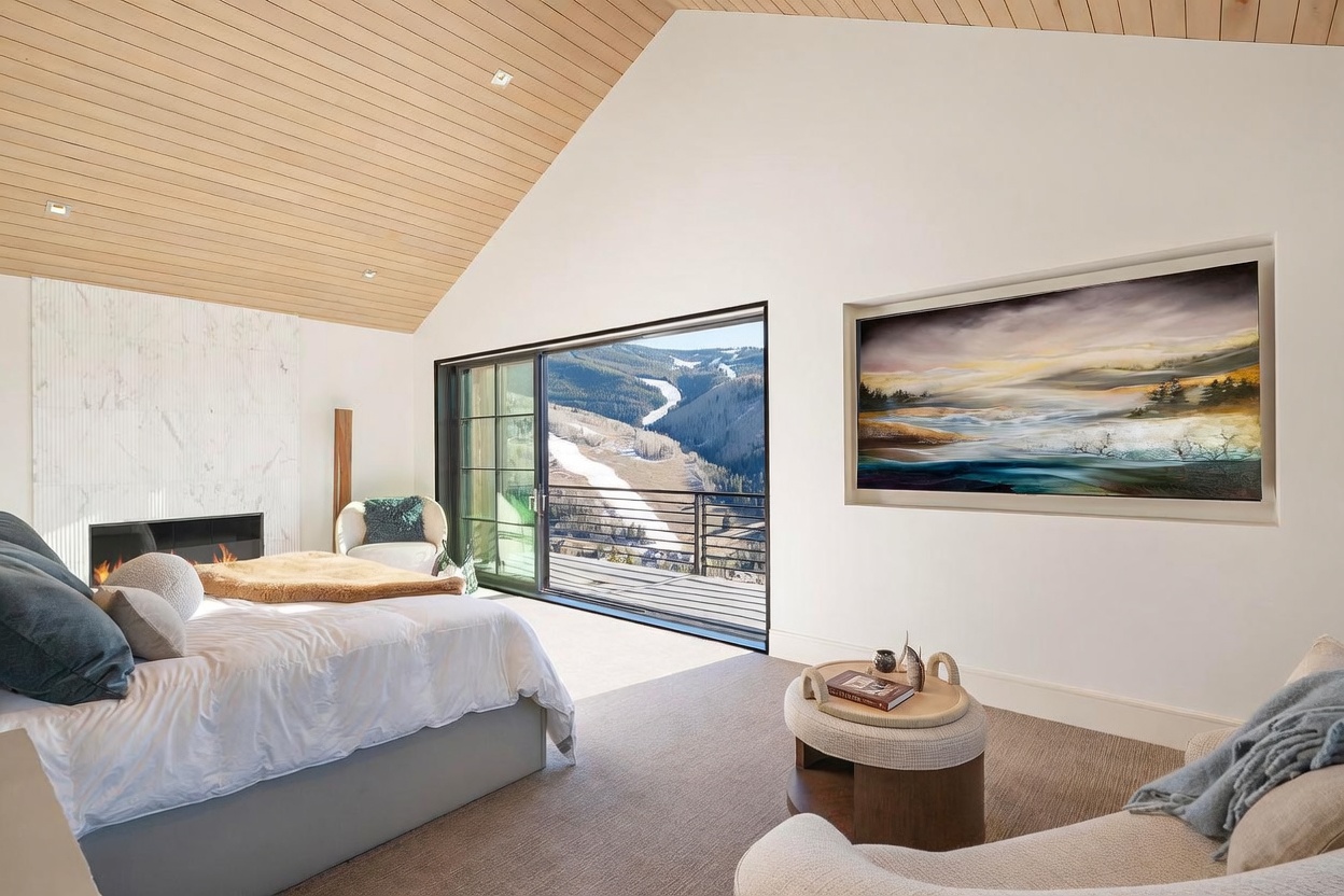 This primary bedroom features a vaulted ceiling with light wood paneling and a large, wall-mounted landscape painting that serves as a focal point. A sliding glass door provides access to a balcony with expansive mountain views, while a stone-clad chimney breast houses a modern fireplace. The room is furnished with a plush bed, a cozy armchair, and a circular ottoman, creating a serene and luxurious atmosphere.