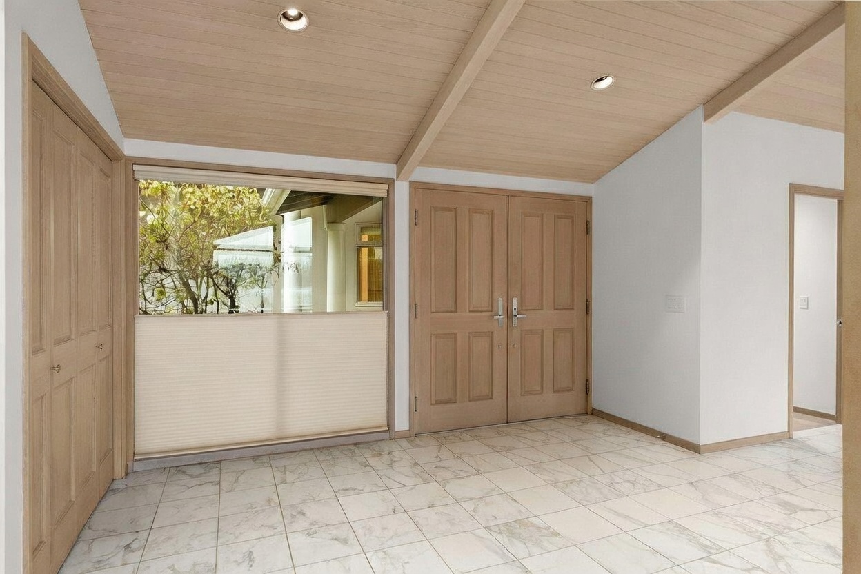This bright entryway features light-toned wood paneling on the vaulted ceiling and matching wood-toned double doors. The floor is covered in large, light-colored tiles with a subtle marble-like pattern, and a large window with a pleated shade allows natural light to filter in. The space has a clean, neutral aesthetic with a simple, open layout that provides a welcoming transition into the home.
