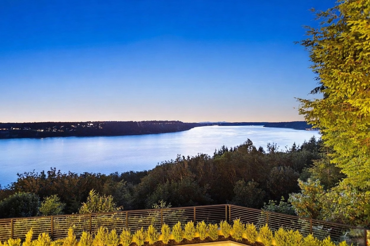 This elevated perspective captures a stunning, expansive water view from a modern balcony featuring a sleek, horizontal-slat metal railing. In the foreground, a row of neatly manicured, vibrant green shrubs lines the edge of the deck, while the background showcases a serene bay framed by dense, dark forest and a clear, twilight sky. The composition emphasizes the property's prime location and tranquil, scenic outlook.
