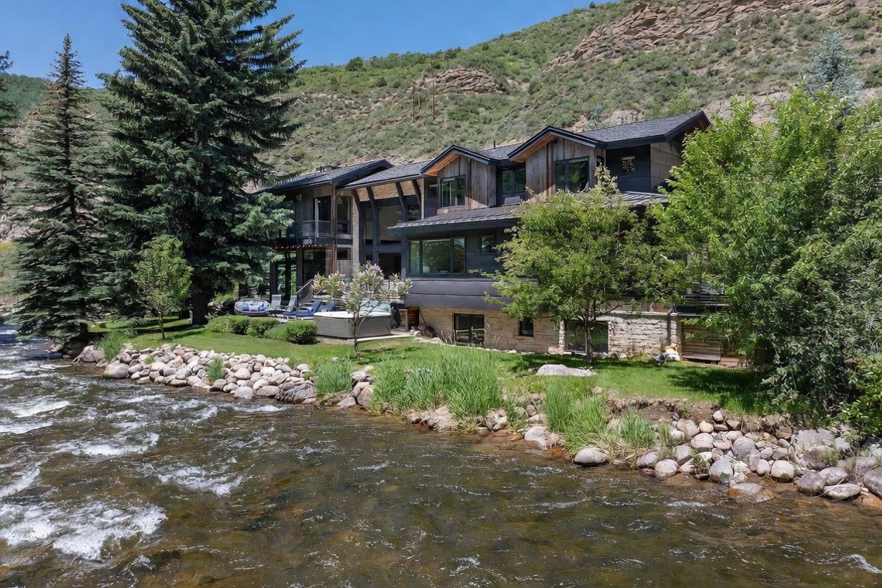 This stunning mountain residence is situated directly along a rushing river, showcasing a multi-level architectural design with a blend of wood siding and stone accents. The rear exterior features a spacious lawn, a private hot tub, and a tiered deck area perfect for outdoor relaxation while enjoying the natural surroundings. The home is framed by mature evergreen trees and a dramatic hillside backdrop, creating a serene and secluded atmosphere.