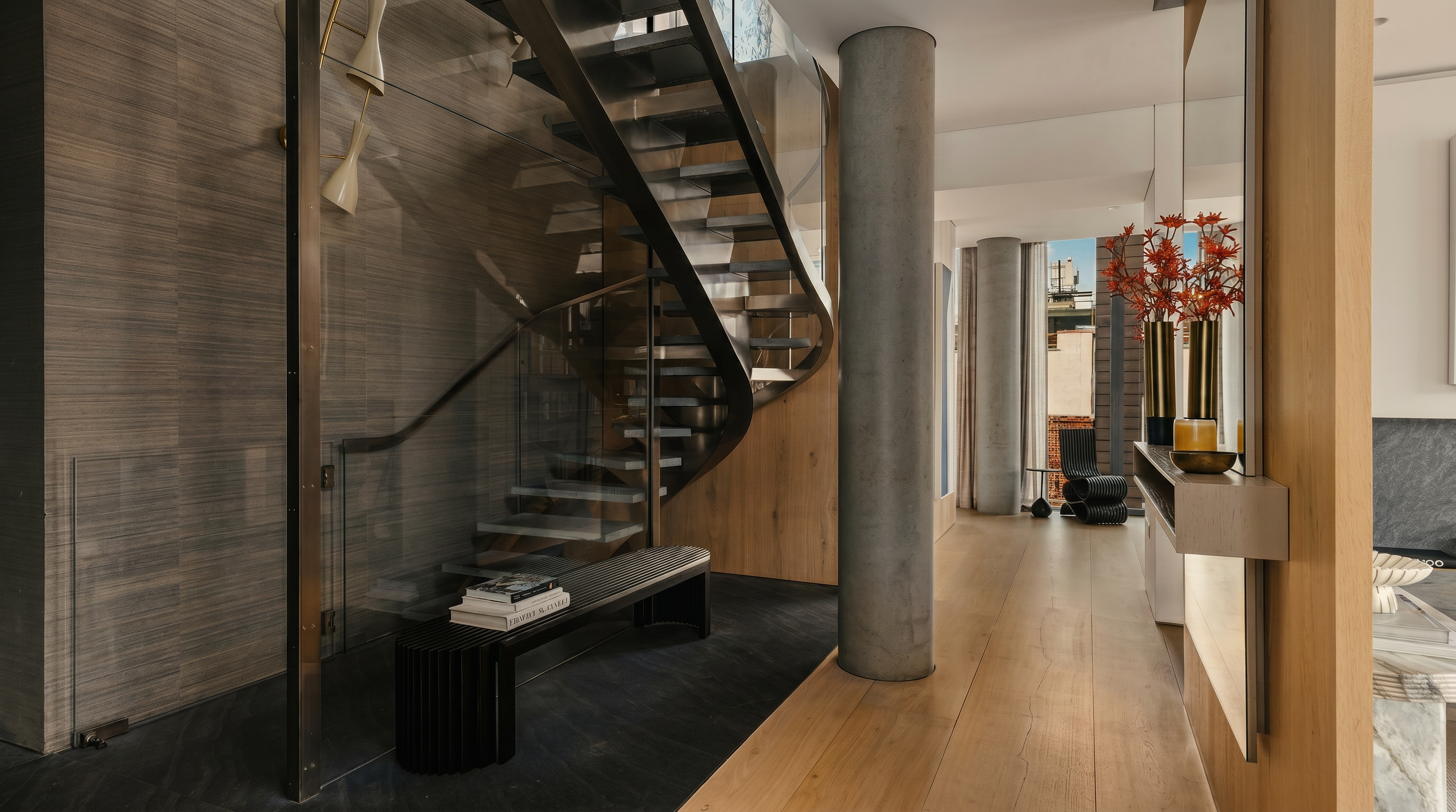 This sophisticated interior space features a striking curved staircase with glass railings and metal treads, serving as a central architectural focal point. The hallway transitions from dark stone flooring near the stairs to light wood planks, leading the eye toward a bright, open area with a concrete column and a minimalist seating arrangement. The design aesthetic is modern and refined, utilizing a neutral color palette and clean lines to create an airy, high-end atmosphere.
