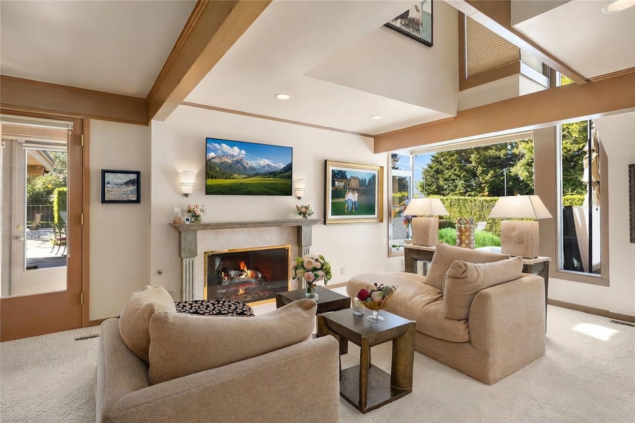 This bright and airy living room features a vaulted ceiling and large windows that provide ample natural light and views of the outdoor greenery. A central fireplace with a stone surround and mantel serves as a focal point, flanked by wall sconces and a mounted television. The space is furnished with comfortable, neutral-toned seating and two small side tables, creating a cozy and inviting atmosphere.