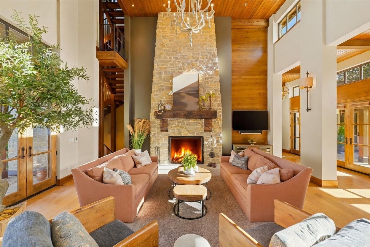This grand living room features a soaring ceiling with exposed wood beams and a prominent stone-clad chimney breast that serves as the central focal point. Two plush, warm-toned sofas face each other across a tiered wooden coffee table, creating an inviting conversation area beneath a large, ornate chandelier. The space is framed by light wood flooring and tall windows, with a staircase visible in the background, contributing to an airy, sophisticated, and rustic-modern aesthetic.
