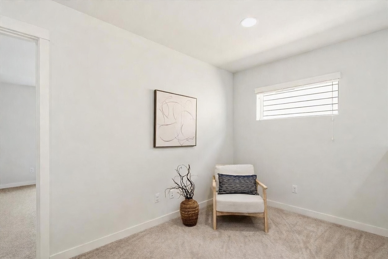 This bright, minimalist corner space features neutral-toned carpeting and light-colored walls, creating a serene atmosphere. A modern armchair with a dark patterned throw pillow sits beside a decorative vase with dried branches, while a piece of abstract line art hangs on the wall above. The room is illuminated by a recessed ceiling light and a horizontal window with blinds, offering a clean and versatile area suitable for a reading nook or home office.