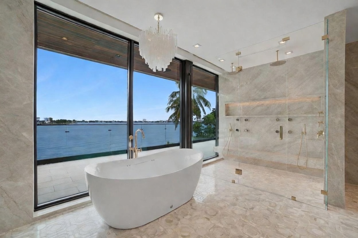 This luxurious primary bathroom features a freestanding soaking tub positioned in front of expansive floor-to-ceiling windows that offer a panoramic view of the water. To the right, a spacious walk-in shower is enclosed by glass panels and finished with large-format stone tiles, including a built-in recessed shelf. The room is illuminated by a decorative crystal chandelier, creating a sophisticated and serene atmosphere.