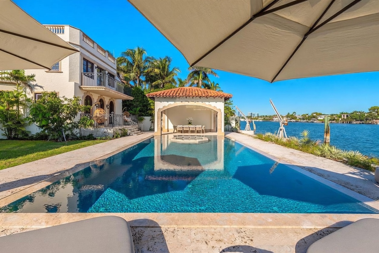 This luxurious waterfront property features a rectangular swimming pool that leads directly toward a covered outdoor pavilion with a dining area. The scene is framed by a large umbrella in the foreground, looking out over the pool toward the calm canal and distant cityscape. The Mediterranean-style architecture of the home, complete with a balcony and palm trees, creates a serene and upscale resort-like atmosphere.