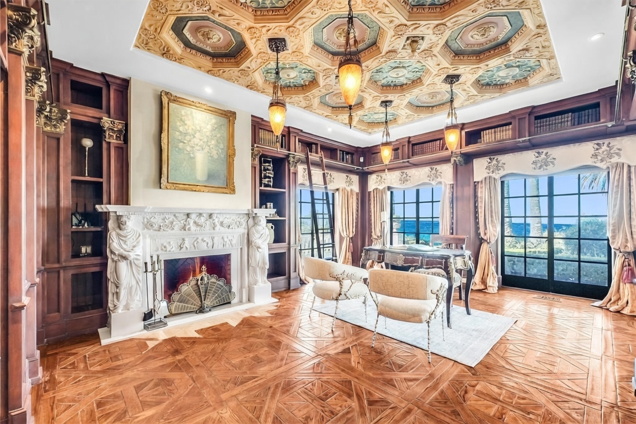 This opulent study features intricate wood paneling, a highly detailed coffered ceiling with decorative medallions, and a grand fireplace with a sculpted stone surround. The room is furnished with a classic desk and two upholstered chairs, while large windows provide a scenic view of the ocean. The space is defined by its rich, traditional aesthetic and sophisticated craftsmanship.