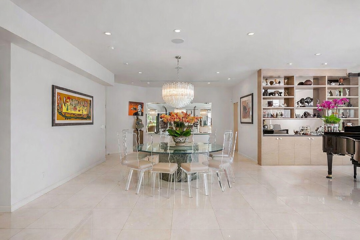 This elegant dining room features a circular glass-topped table surrounded by six modern, transparent chairs, positioned beneath a large, multi-tiered crystal chandelier. To the right, a built-in light wood shelving unit displays decorative objects and art, while a grand piano sits partially in view. The space is finished with light-colored tile flooring and neutral walls, creating a bright and sophisticated atmosphere.
