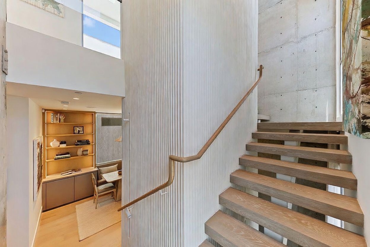 This interior view captures a modern staircase with light wood treads and a sleek, minimalist handrail. To the left, a textured, vertical-slatted wall leads the eye toward a cozy, recessed study area featuring built-in shelving and a small dining table. The space is characterized by clean lines, a neutral color palette, and a blend of raw concrete and warm wood textures.