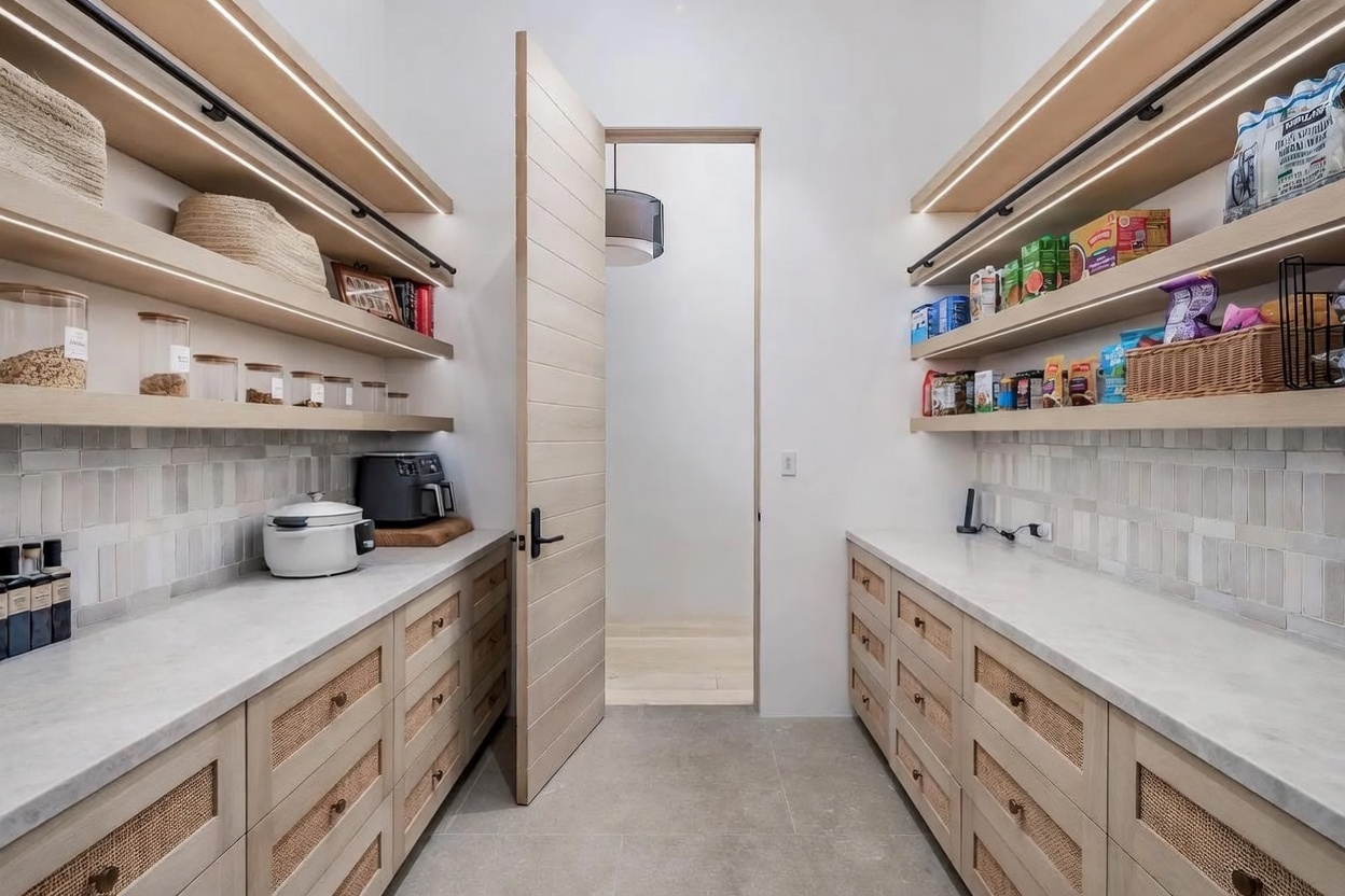 This organized walk-in pantry features light wood cabinetry with woven cane detailing and stone countertops. Open shelving with integrated under-shelf lighting provides ample storage for dry goods and kitchen essentials, while a central door leads to an adjacent room. The space is finished with a neutral tile backsplash and large-format floor tiles, creating a clean and modern aesthetic.