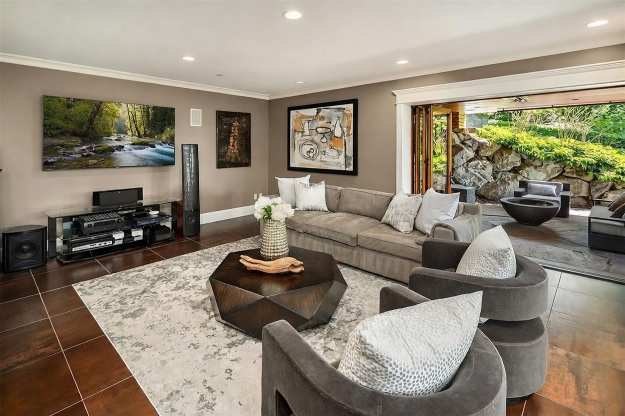 This sophisticated living room features a large, plush gray sofa and two matching rounded armchairs arranged around a dark, geometric coffee table on a textured area rug. The space is anchored by a large wall-mounted television and a media console, while a wide, folding glass door system seamlessly connects the interior to an outdoor patio area with a fire pit. The room is finished with dark tile flooring and neutral wall tones, creating a cohesive and inviting atmosphere for relaxation or entertaining.