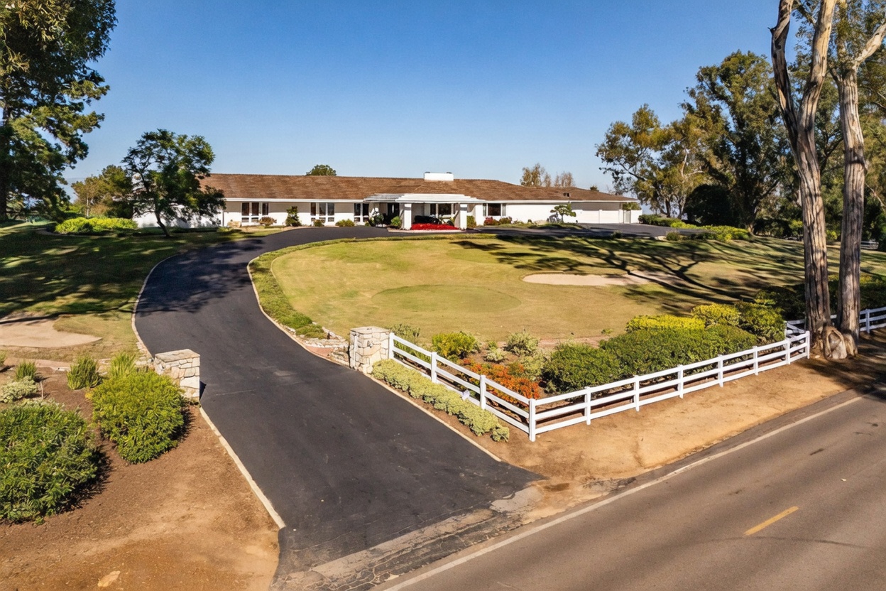 This elevated perspective captures a sprawling single-story ranch-style home set back from the road behind a white split-rail fence. A long, curved asphalt driveway leads up to the main entrance, which is framed by a covered portico and surrounded by a manicured lawn featuring a small sand bunker. The property is framed by mature trees and lush landscaping, creating a private and expansive estate-like atmosphere.