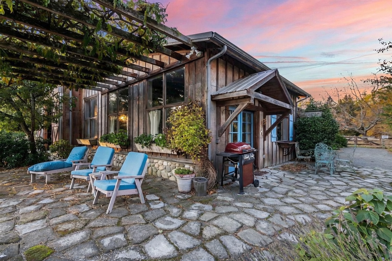 This rustic, wood-sided structure features a charming stone-paved patio shaded by a wooden pergola draped in climbing vines. The space is furnished with blue lounge chairs and a grill, creating an inviting outdoor living area set against a vibrant sunset sky. The architectural style emphasizes natural textures and a cozy, countryside aesthetic.