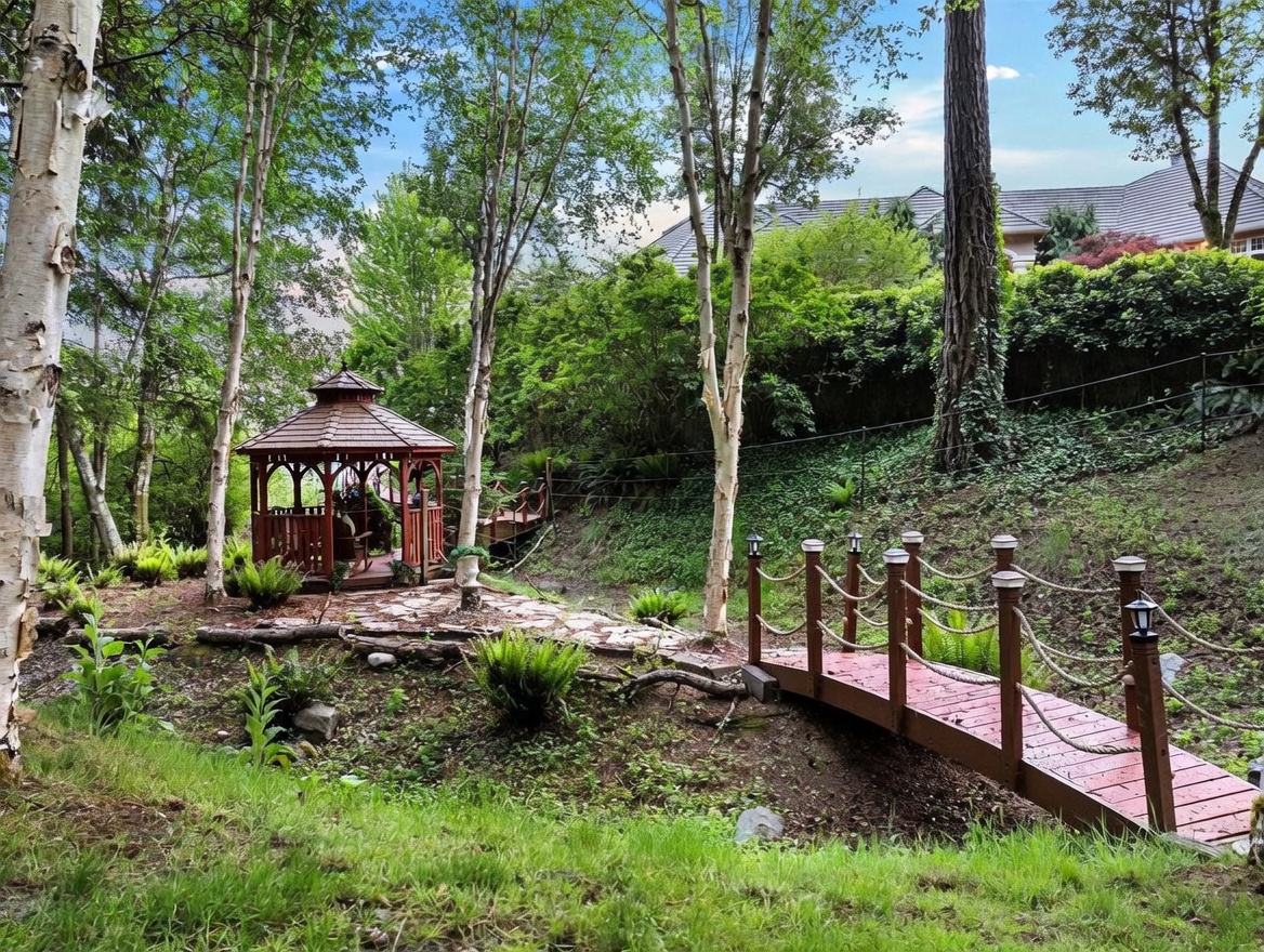 This serene backyard garden features a wooden gazebo nestled among mature trees and lush greenery. A small, decorative wooden footbridge with rope railings spans a dry creek bed, leading toward the gazebo along a stone path. The scene offers a tranquil, park-like atmosphere with a gentle slope and dense landscaping, providing a private and peaceful outdoor retreat.