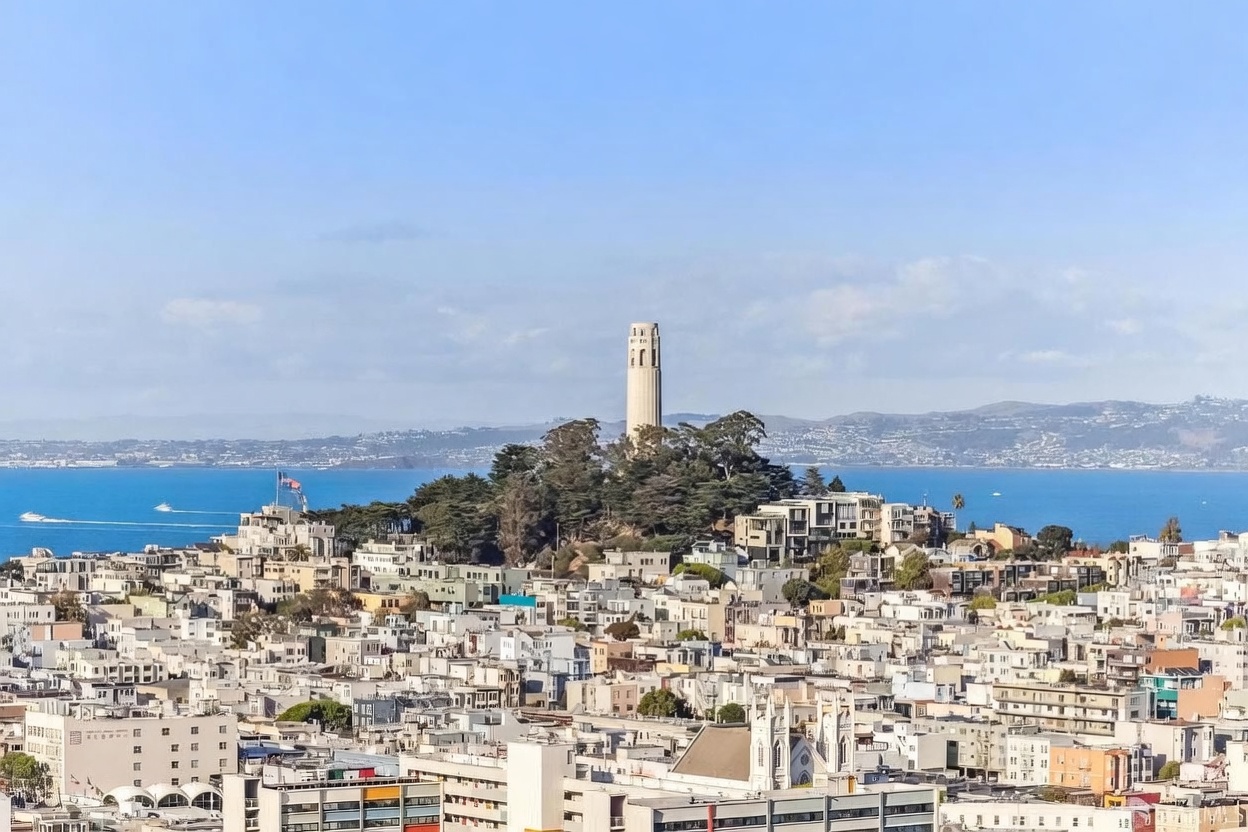 This high-angle aerial view captures a dense urban landscape in San Francisco, prominently featuring the iconic Coit Tower perched atop Telegraph Hill. The scene showcases a sprawling cityscape of residential buildings and historic architecture, with the expansive blue waters of the San Francisco Bay and distant hills providing a scenic backdrop. The perspective offers a cinematic overview of the neighborhood's density and its proximity to the waterfront.