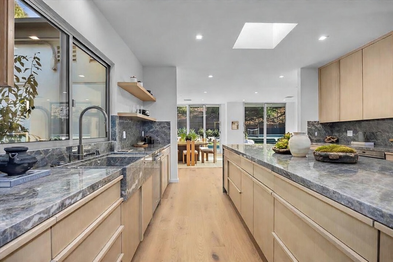 This modern kitchen features light wood cabinetry paired with dark, veined stone countertops and a matching backsplash. A large central island provides ample workspace, while a deep sink is positioned beneath a wide window that overlooks the outdoor dining area. The open-concept design flows seamlessly into the adjacent dining space, enhanced by recessed lighting and a skylight that brightens the room.