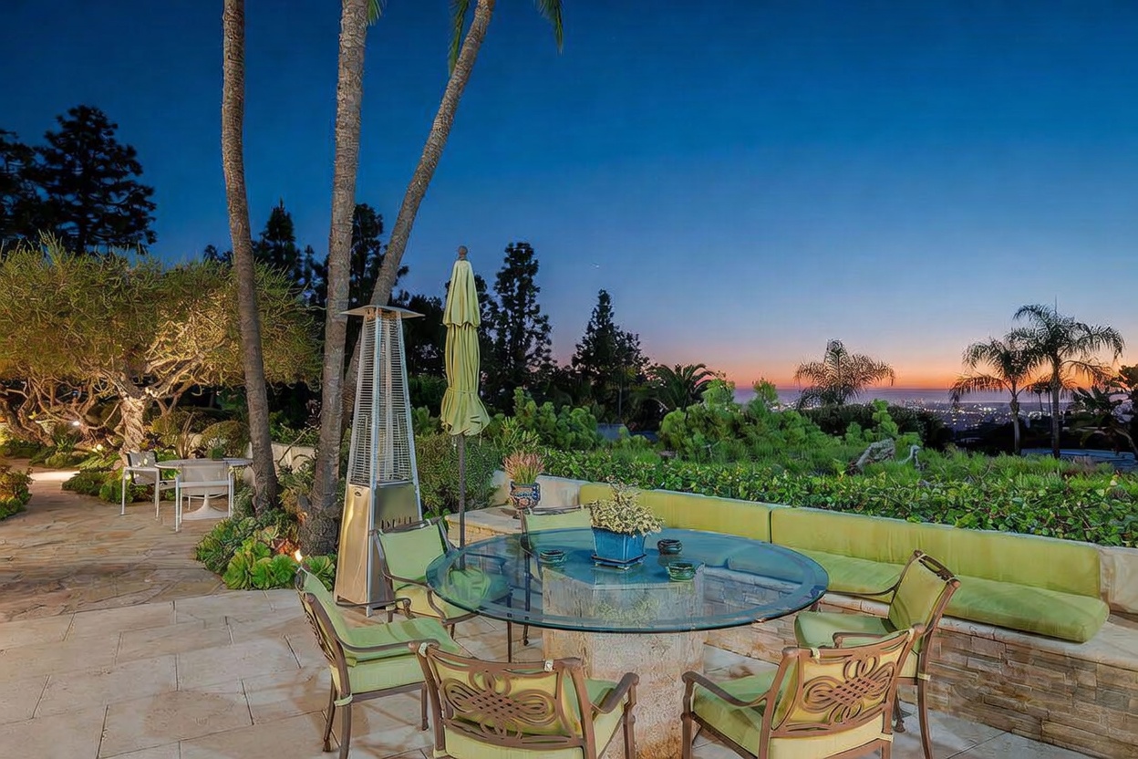 This outdoor patio space features a glass-topped dining table surrounded by cushioned chairs, set against a backdrop of lush greenery and a panoramic sunset view. A tall metal patio heater stands nearby, and a long built-in bench with green cushions provides additional seating along the stone wall. The scene is captured at dusk, highlighting the warm ambient lighting and the expansive, scenic horizon.