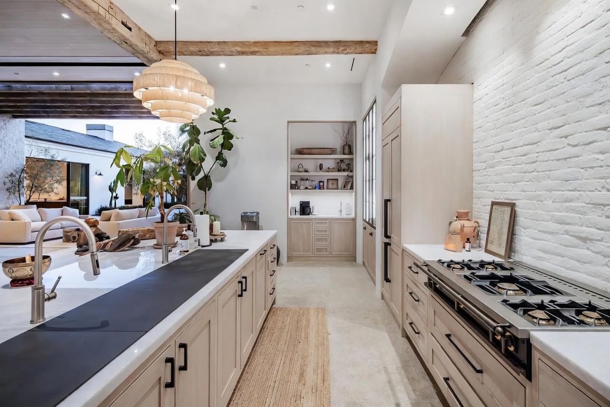 This bright, open-concept kitchen features light wood cabinetry, a large island with a dark stone prep area, and a professional-grade range. A white brick wall adds texture to the space, while a built-in shelving unit provides an elegant display area. The perspective looks through the kitchen toward an adjacent living space, highlighting the seamless flow between rooms.