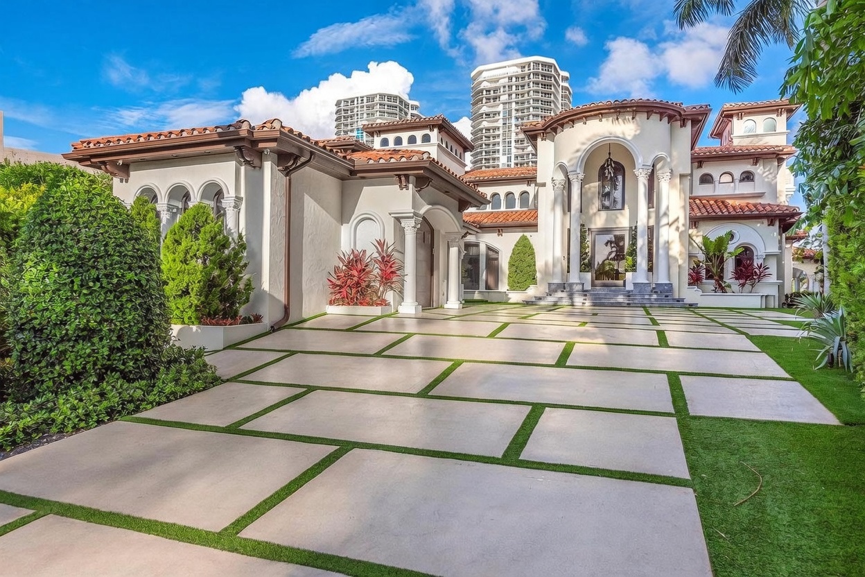 This grand Mediterranean-style estate features a prominent arched entryway with decorative columns and a multi-level roofline with terracotta tiles. The expansive driveway is composed of large concrete pavers separated by strips of manicured grass, leading up to the impressive facade. Lush landscaping, including mature shrubs and palm trees, frames the home, while a clear blue sky with scattered clouds provides a bright, inviting backdrop.