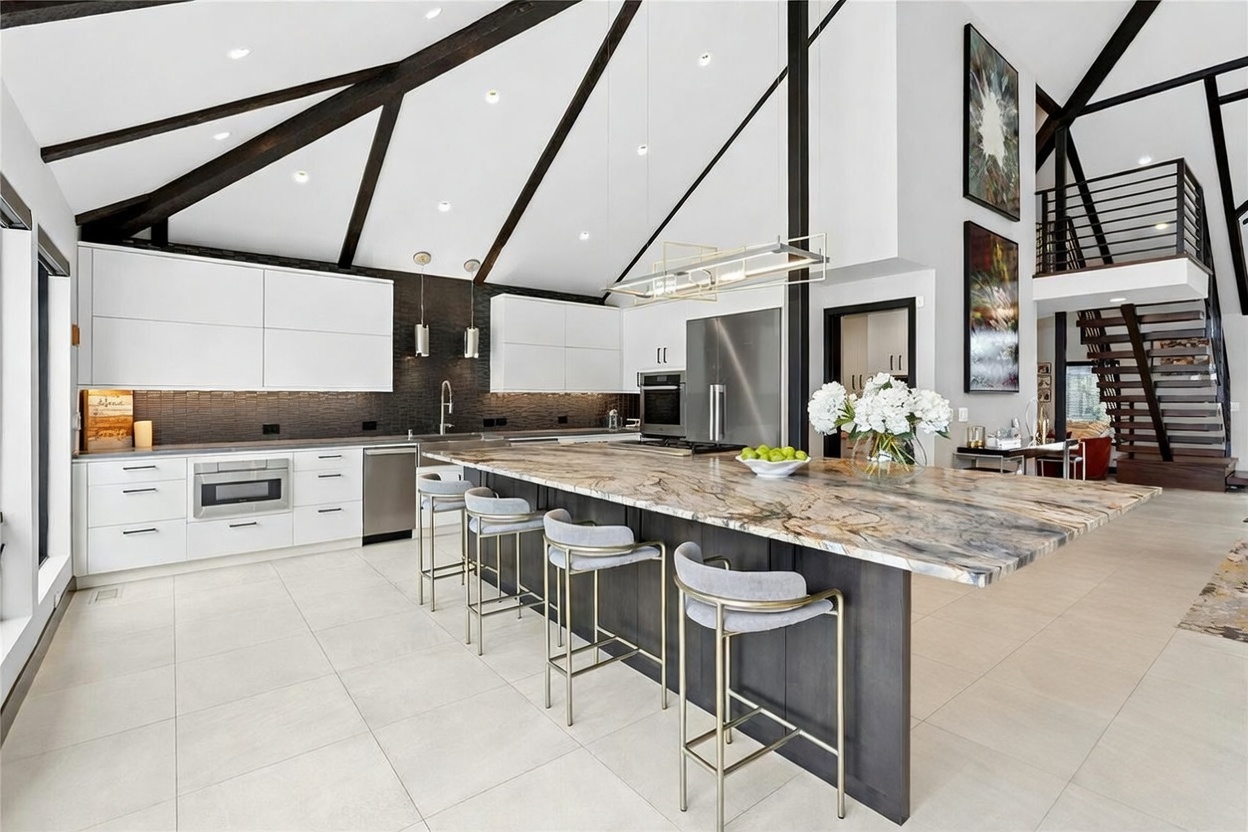 This stunning, high-end kitchen features a dramatic vaulted ceiling with exposed dark wood beams and a massive, waterfall-edge stone island that serves as the centerpiece. The space is defined by a sophisticated contrast between sleek white upper cabinetry, dark lower cabinets, and a textured dark backsplash, all illuminated by modern linear pendant lighting. The open-concept design seamlessly integrates with a staircase in the background, creating an airy, luxurious atmosphere perfect for entertaining.