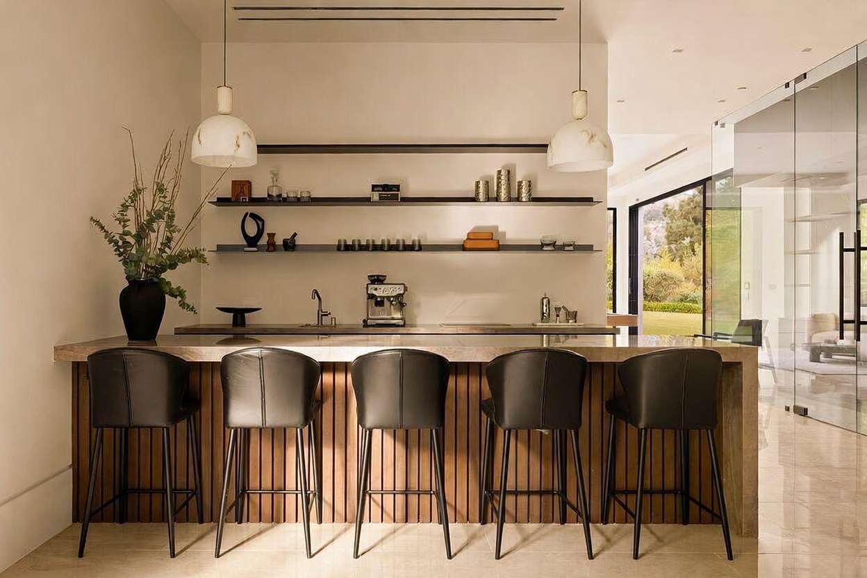 This modern kitchen bar area features a long stone-topped counter with five sleek, dark-upholstered stools set against a vertical wood-slatted base. Behind the counter, open shelving displays curated decor, while a professional-grade espresso machine sits on the workspace. The space is illuminated by two elegant pendant lights, and a large glass wall to the right provides a bright, open view of the outdoor landscape.