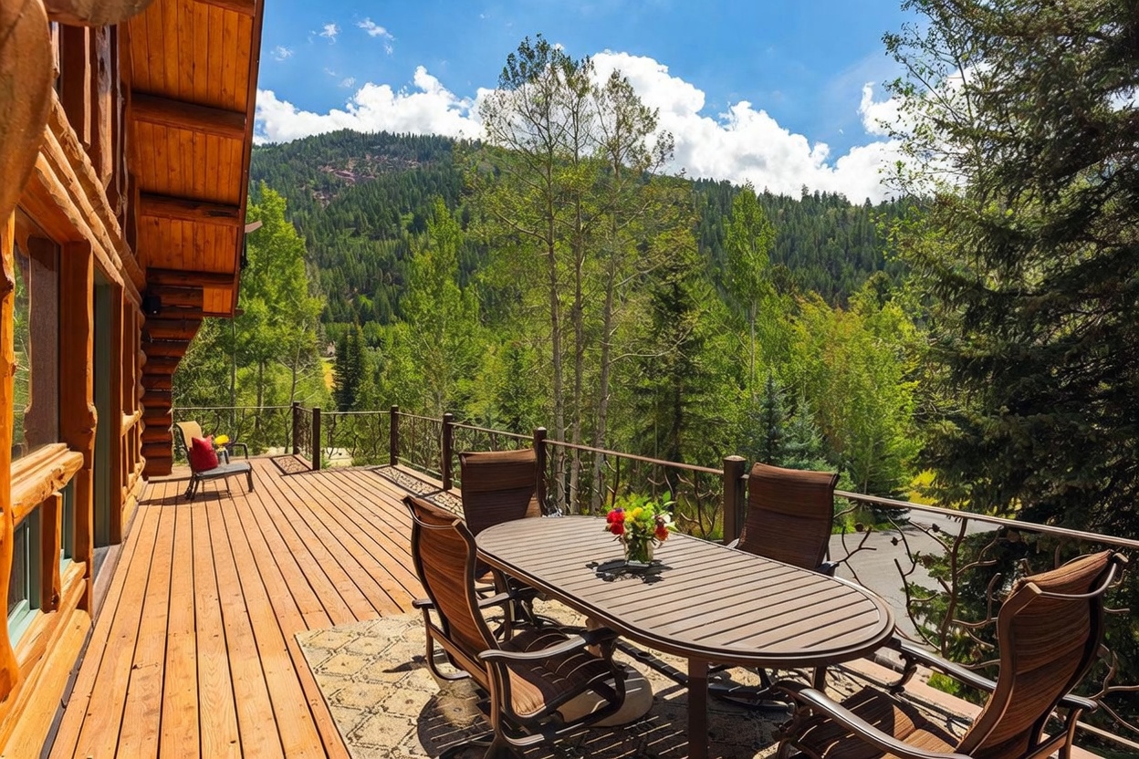 This expansive wooden deck offers a serene outdoor living space with a dining set and additional seating, perfect for enjoying the surrounding mountain views. The deck features a decorative metal railing and overlooks a lush forest of aspen and pine trees under a bright, partly cloudy sky. The rustic log construction of the home's exterior adds to the warm, inviting atmosphere of this elevated vantage point.