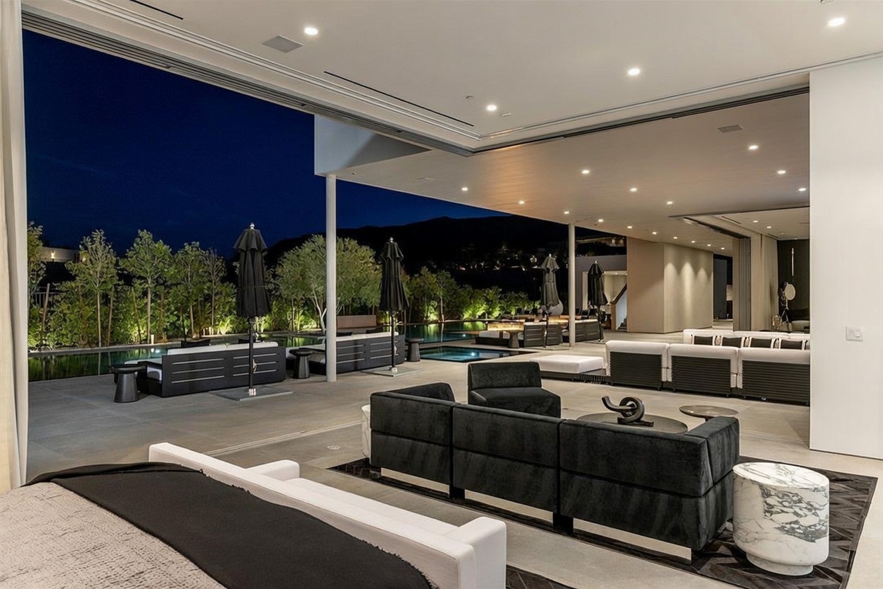 This expansive indoor-outdoor living space features a seamless transition from a modern interior lounge to a covered patio overlooking a swimming pool at night. The area is furnished with multiple dark-toned modular seating arrangements, sleek side tables, and large umbrellas, all set against a backdrop of illuminated landscaping and distant hills. The open-concept design emphasizes a luxurious, resort-style atmosphere perfect for evening entertaining.