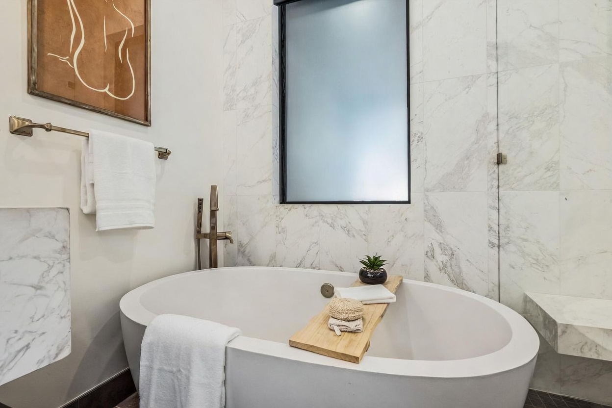 This elegant bathroom features a freestanding white soaking tub as the centerpiece, complemented by a wooden bath tray holding a small potted plant and bath accessories. The wall behind the tub is finished with large-format stone tiles with prominent veining, and a tall, frosted window provides natural light while maintaining privacy. A brass-toned wall-mounted faucet and a matching towel rack add a touch of warmth to the sophisticated, neutral-toned space.