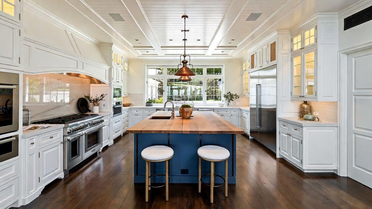 This bright and spacious kitchen features a large central island with a wood countertop and a deep blue base, complemented by crisp white cabinetry and stainless steel appliances. The room is anchored by a coffered ceiling with recessed lighting and a prominent pendant light fixture, while large windows above the sink provide a clear view of the outdoors. The dark wood flooring creates a sophisticated contrast against the light-toned walls and cabinetry, offering a clean and high-end aesthetic.