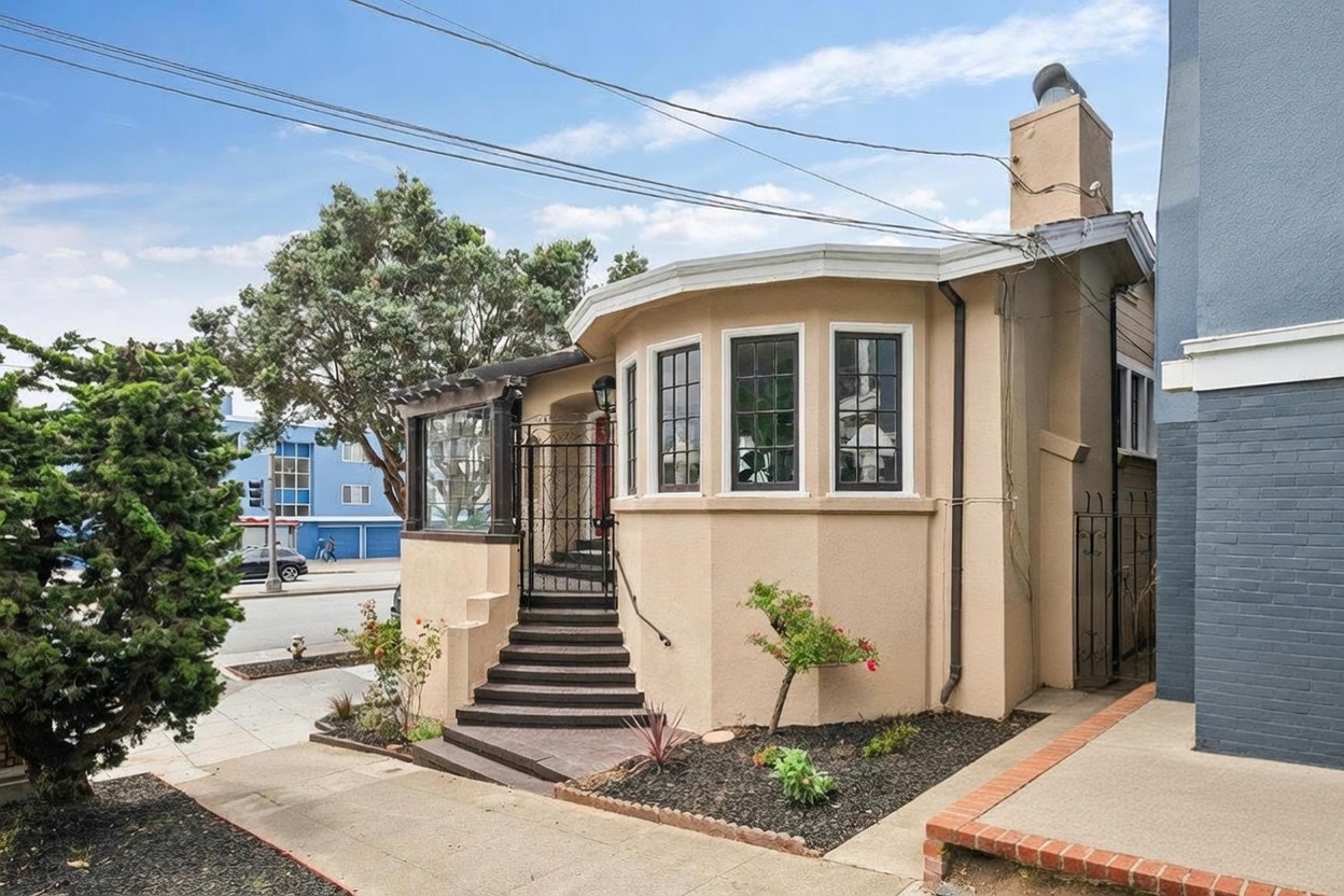 This charming single-story home features a distinctive curved bay window facade and a prominent chimney on the side. A set of dark-toned stairs leads up to the gated entryway, which is framed by a decorative pergola structure. The exterior is finished in a warm, neutral stucco, complemented by a well-maintained front garden with a large bush and mature trees.