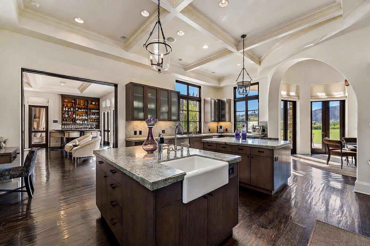 This spacious, high-end kitchen features two large islands with stone countertops and dark cabinetry, centered under a coffered ceiling with pendant lighting. The room is anchored by dark wood flooring and opens into an adjacent bar area, with large windows and glass doors providing natural light and views of the outdoors. The overall aesthetic is sophisticated and traditional, emphasizing a grand, open-concept layout suitable for entertaining.