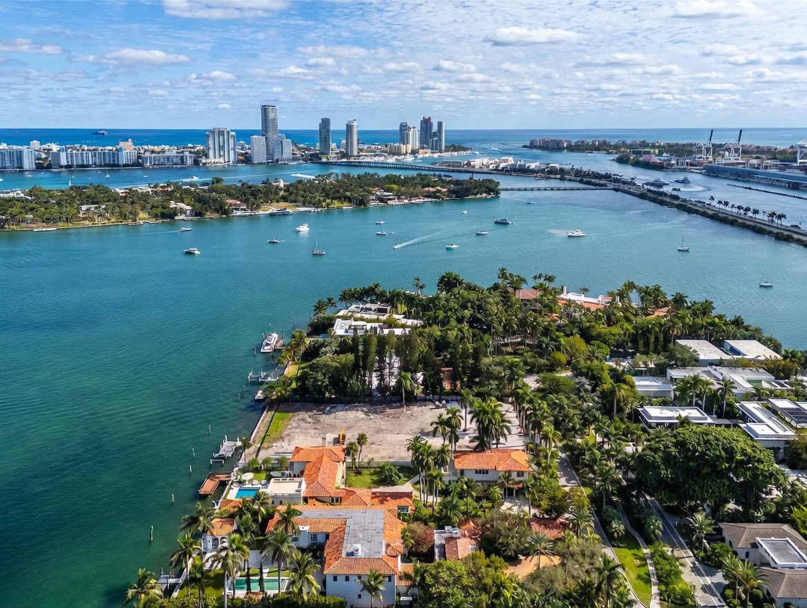 This high-angle aerial view captures a sprawling waterfront estate situated on a lush, tropical peninsula. The property features a large, cleared building site, mature palm trees, and a multi-winged residence with traditional terracotta-tiled roofs and a private swimming pool. In the background, the expansive blue bay stretches toward a distant city skyline, creating a serene and prestigious coastal atmosphere.
