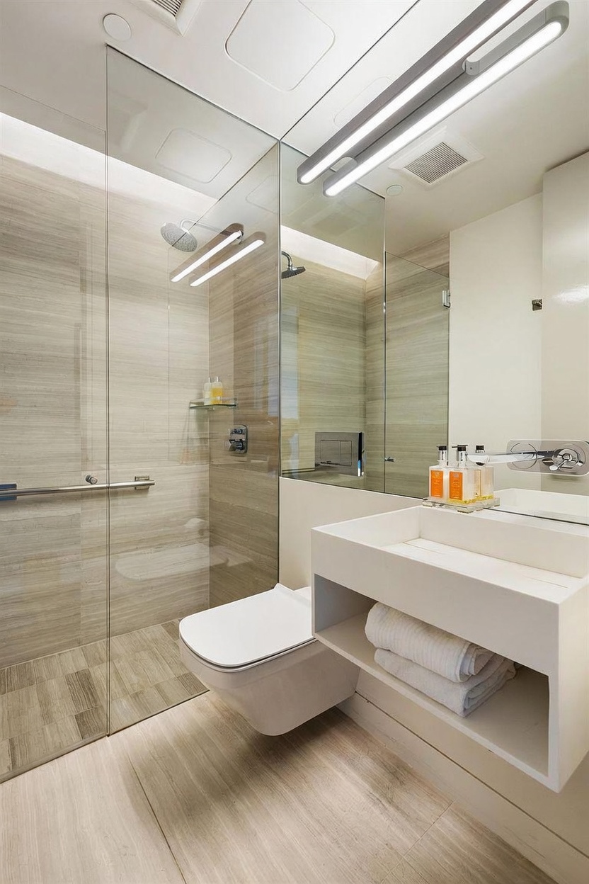 This modern, minimalist bathroom features a sleek wall-mounted vanity with integrated shelving and a glass-enclosed walk-in shower. The space is finished with light-toned, linear-patterned tile on both the walls and floor, creating a cohesive and airy aesthetic. A wall-mounted toilet and contemporary fixtures complete the clean, high-end design of this compact yet functional space.