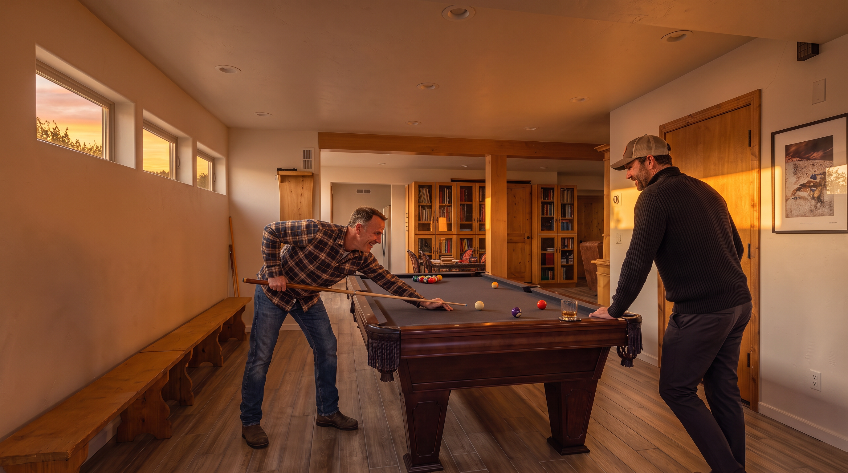 This spacious basement recreation area features a prominent pool table placed centrally on wood-look tile flooring. The space is accented by natural wood elements, including built-in bookshelves, a long bench, and doors, creating a warm and functional atmosphere perfect for entertainment. The clean, neutral color palette and recessed lighting enhance the open layout of this versatile living space.