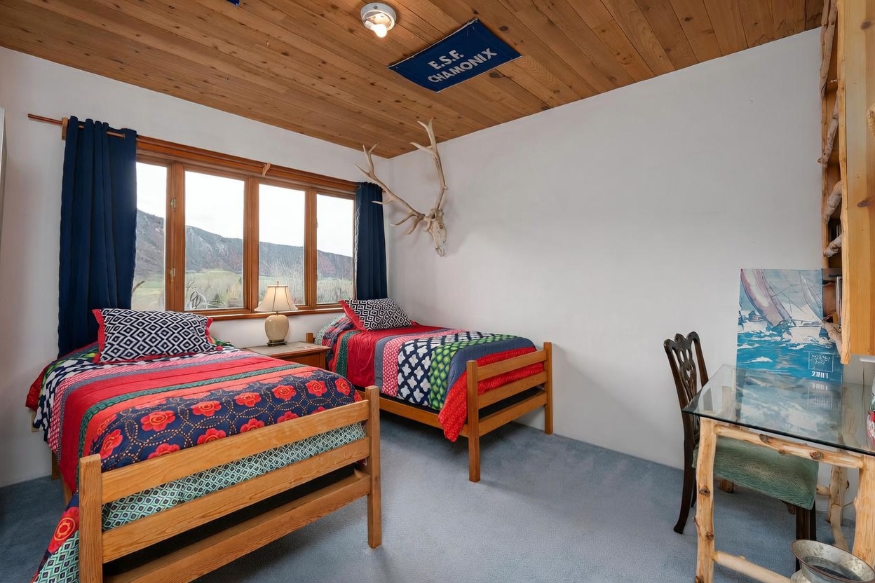 This guest bedroom features two twin beds with vibrant, patterned quilts and a shared nightstand with a lamp. The room is accented by a wood-paneled ceiling, a decorative antler mount on the wall, and a small glass-topped desk area. Large windows provide natural light and a scenic view of the mountains, creating a cozy and rustic atmosphere.