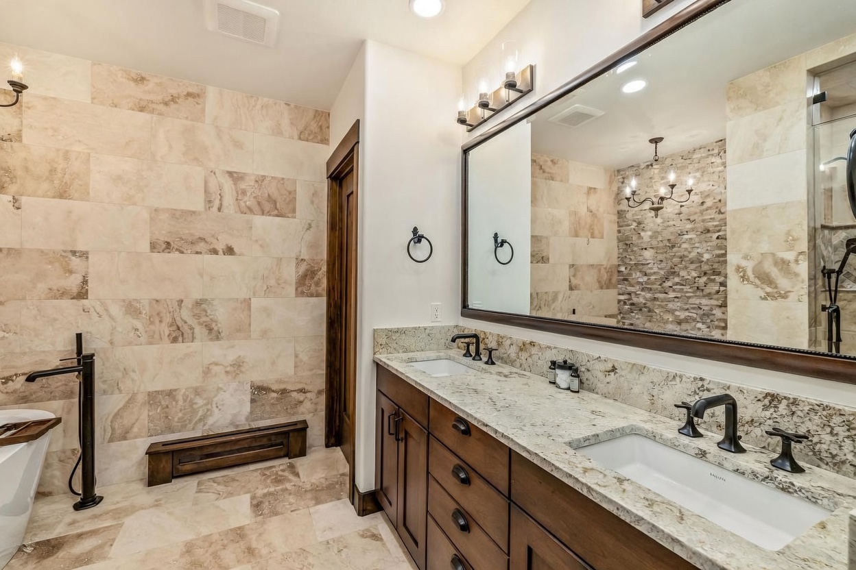 This primary bathroom features a double vanity with a light-toned stone countertop and dark wood cabinetry, complemented by a large mirror reflecting a stone-accented wall. The space is finished with neutral-toned wall and floor tiles, creating a cohesive and elegant aesthetic. A dark-finished faucet and hardware provide a sharp contrast against the lighter surfaces, while a chandelier adds a touch of sophistication to the room.