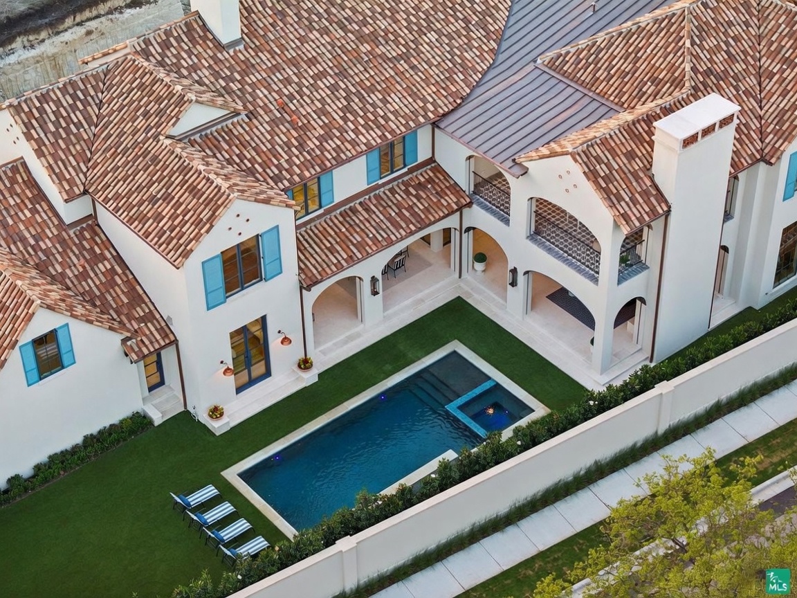 This high-angle aerial perspective captures a luxurious Mediterranean-style residence featuring a terracotta tile roof and crisp white stucco walls. The property includes a private courtyard with a rectangular swimming pool, a spa, and a manicured lawn bordered by a privacy wall. The architectural design emphasizes indoor-outdoor living with covered loggias and balconies that overlook the serene pool area.