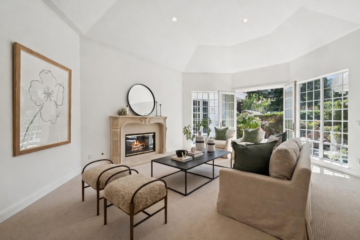This elegant and bright living room features a sophisticated cream-toned sofa, two textured ottomans, and a contemporary black coffee table arranged around a central fireplace. A large, minimalist line-art print hangs on the wall, while expansive windows and glass doors provide abundant natural light and a seamless connection to the outdoor space. The high, vaulted ceiling and neutral color palette create an airy, serene, and inviting atmosphere.