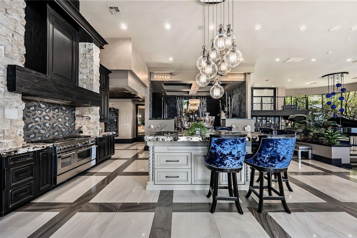 This expansive, high-end kitchen features a large central island with dark stone countertops and vibrant blue velvet bar stools. The space is anchored by a prominent stone-clad chimney breast above the range and a striking multi-globe chandelier hanging over the island. The room showcases a sophisticated design with contrasting light and dark cabinetry, polished tile flooring, and an open-concept layout that flows into a secondary living or dining area.