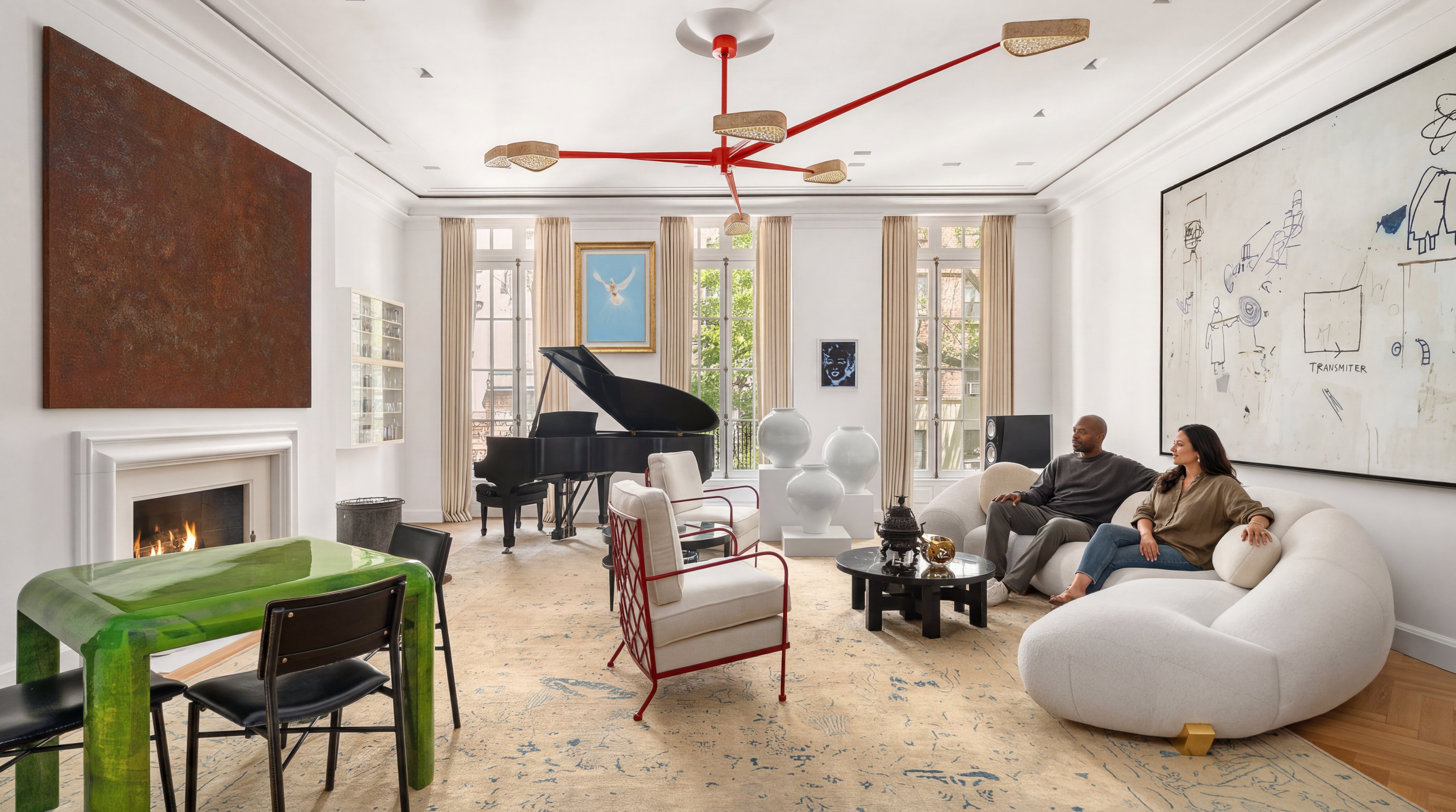 This sophisticated, expansive living room features a blend of contemporary art and classic elements, including a grand piano and a striking red chandelier. The space is anchored by a large, ivory-toned conversation-style sofa and patterned area rug, set against a backdrop of floor-to-ceiling windows and large-scale fine art pieces. Its airy, open-concept design creates an impressive, gallery-like atmosphere perfect for both high-end entertaining and relaxation.
