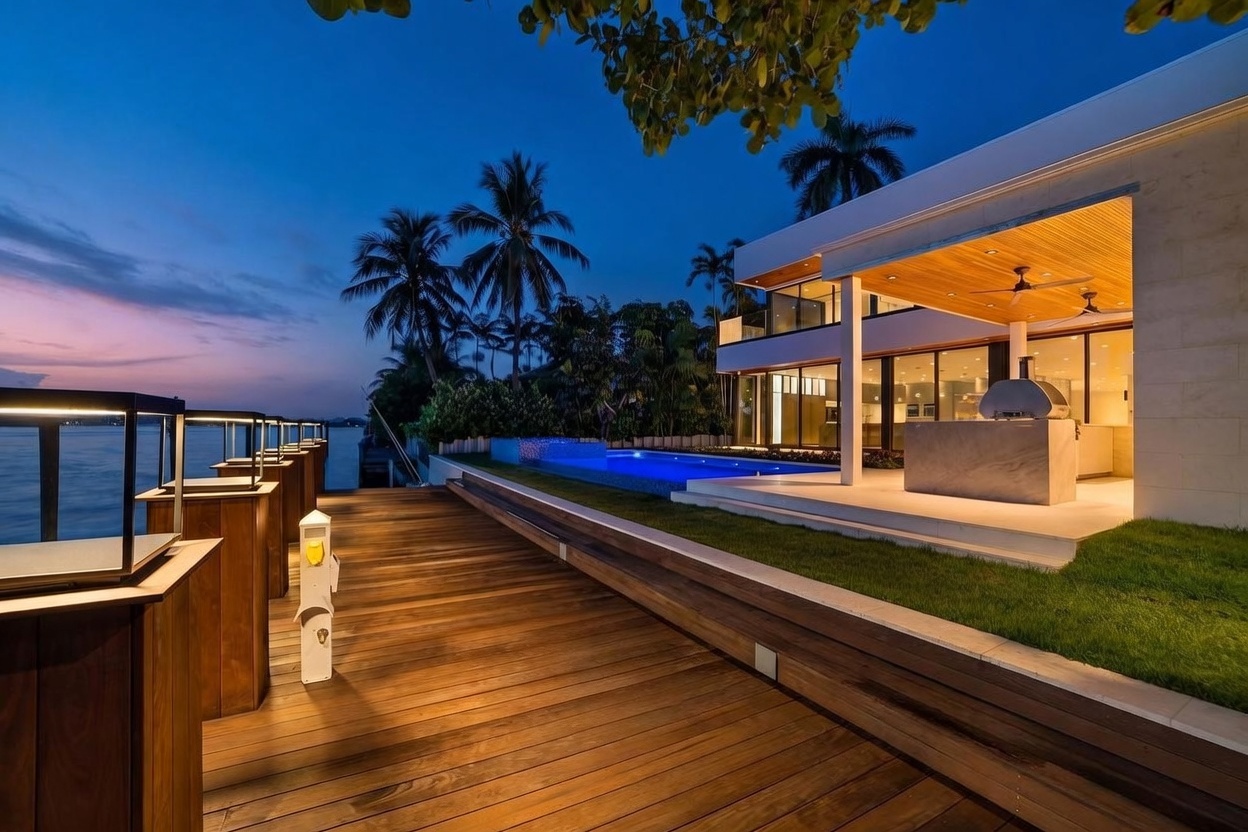 This stunning waterfront property features a modern architectural design with a spacious wooden deck and a glowing blue infinity-edge pool at twilight. The home includes a covered outdoor kitchen area with a stone pizza oven and warm wood-paneled ceilings, seamlessly blending indoor and outdoor living. The scene is captured from a low-angle perspective on the dock, emphasizing the expansive water views and the sophisticated, resort-like atmosphere of the residence.