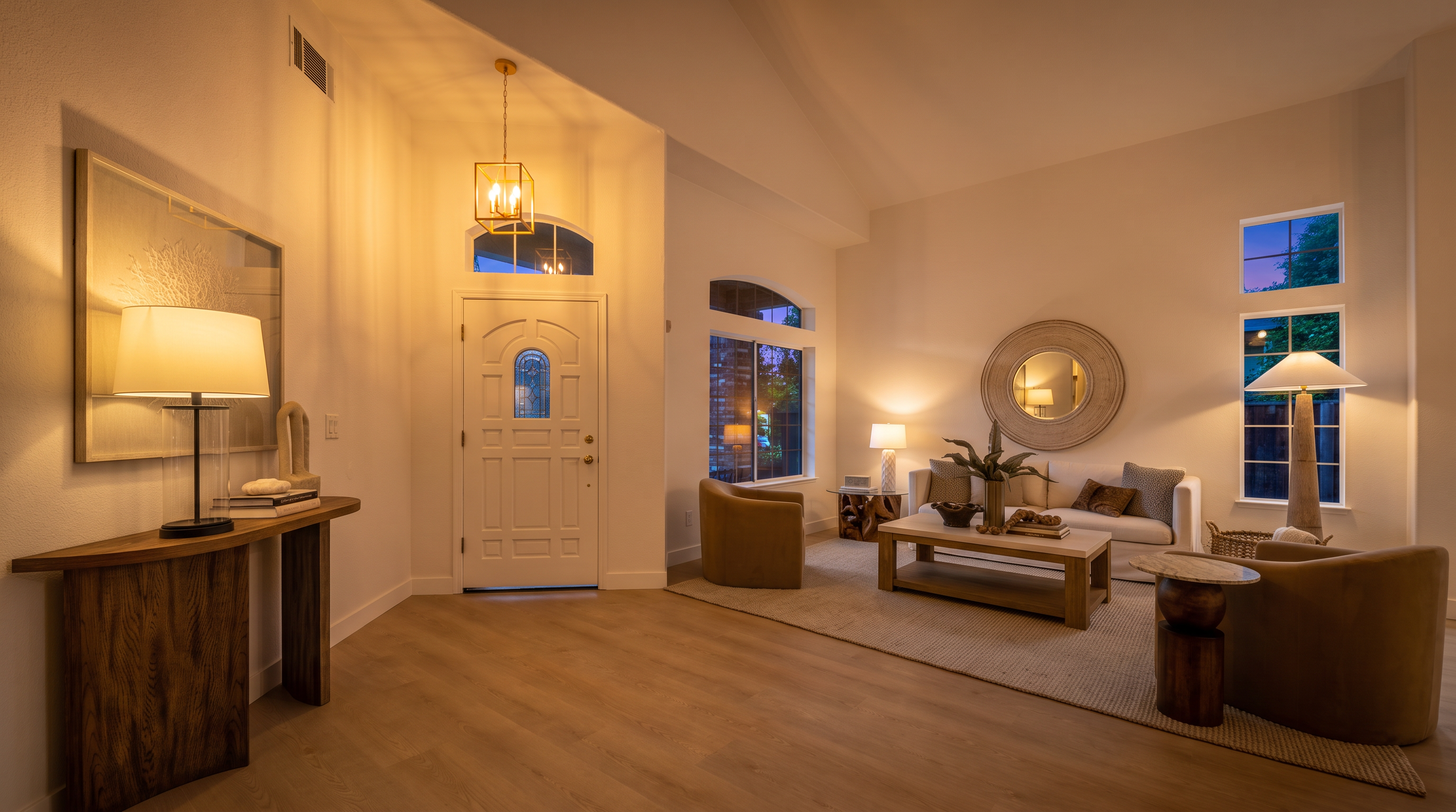 This bright and airy living room features a vaulted ceiling and light wood-toned flooring, creating an inviting open-concept atmosphere. The space is furnished with a neutral sofa, two curved armchairs, and a central coffee table, all arranged on a textured area rug. A console table with a lamp sits in the foreground, while large windows and a front door with a glass insert allow for ample natural light to fill the room.