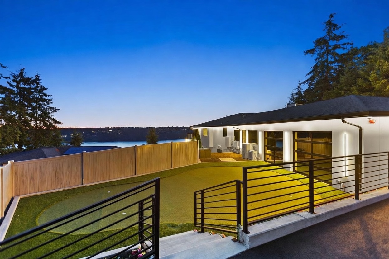 This exterior view captures a modern backyard featuring a synthetic turf putting green enclosed by a tall privacy fence. The house showcases a contemporary design with large glass garage doors and integrated exterior lighting, set against a scenic backdrop of a body of water at dusk. A concrete staircase with black metal railings leads down to the yard, emphasizing a clean, structured landscape design.