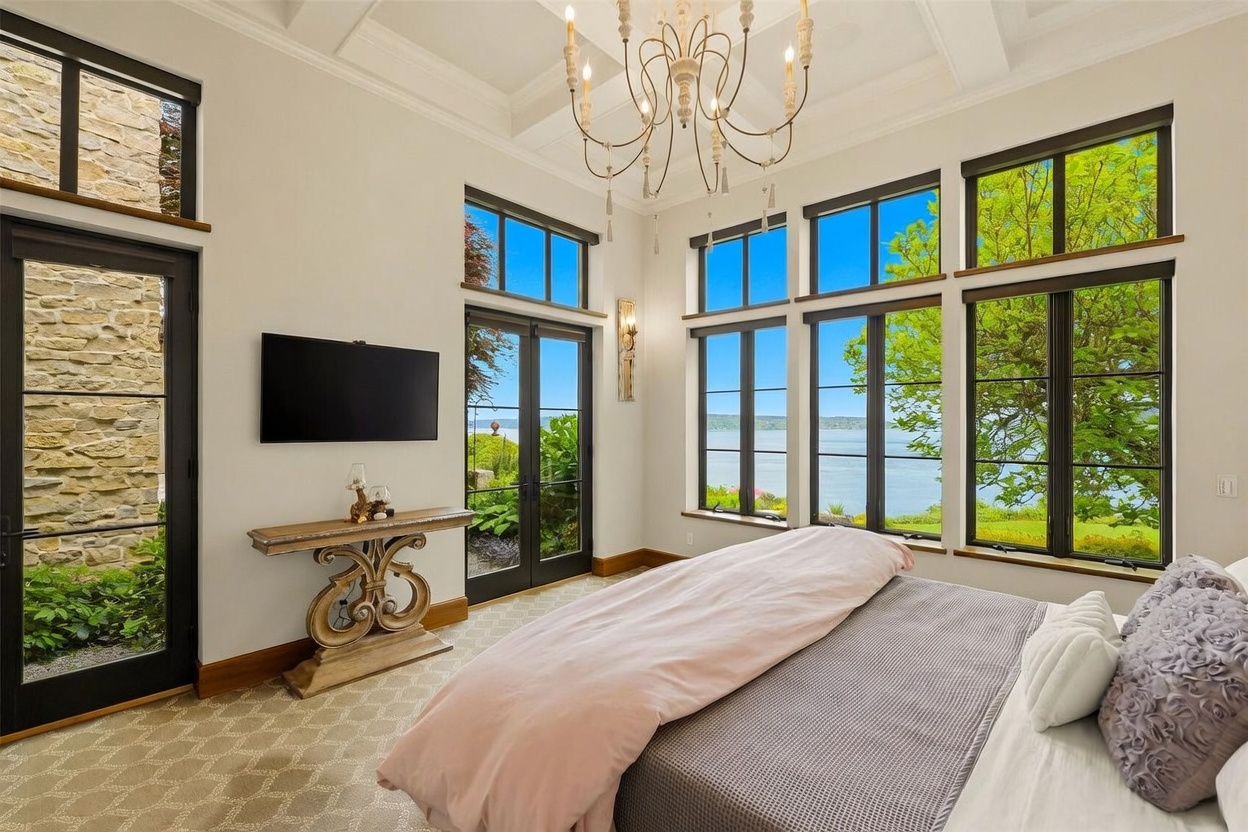This elegant primary bedroom features a high coffered ceiling with a decorative chandelier and expansive windows that offer a panoramic view of the water. The room is furnished with a bed dressed in soft pink and grey linens, complemented by a console table placed against a wall with a mounted television. The space is bright and airy, with a neutral color palette and a stone-clad wall section near the glass door, creating a sophisticated and tranquil atmosphere.