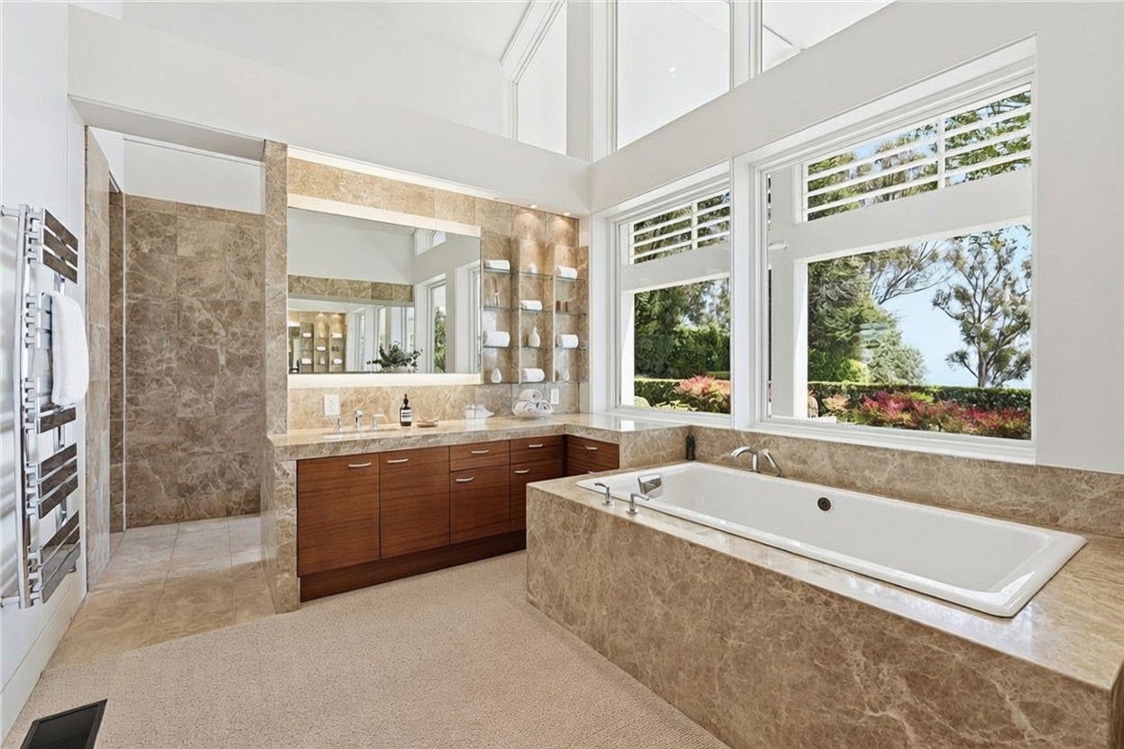 This spacious primary bathroom features a large soaking tub encased in stone, positioned next to expansive windows that provide ample natural light and a view of the outdoors. A long vanity with wood-toned cabinetry and a large mirror sits against a stone-tiled wall, complemented by open shelving for storage. The room is characterized by its high ceilings and a neutral, sophisticated color palette, creating a bright and airy atmosphere.