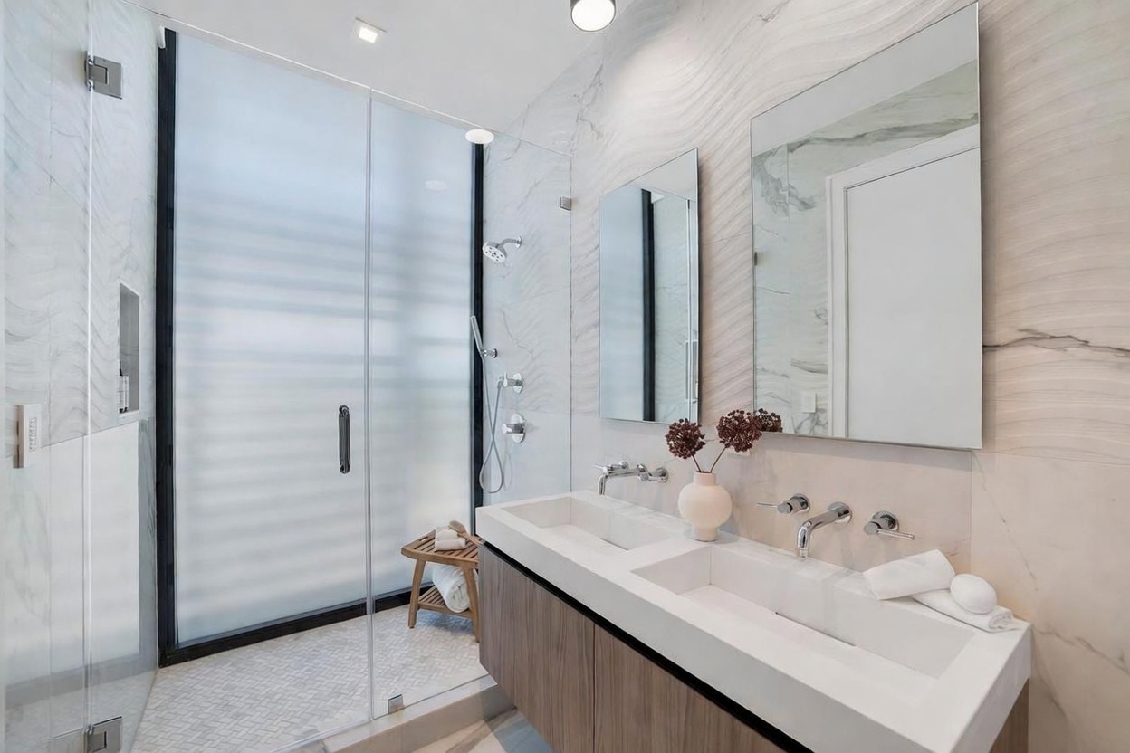 This modern primary bathroom features a sleek double vanity with a light wood-toned cabinet and a long, integrated dual-basin sink. To the left, a spacious walk-in shower is enclosed by glass panels and features a frosted glass door, a handheld showerhead, and a small wooden stool. The walls are finished with large-format, light-toned stone tiles with subtle veining, complemented by two rectangular mirrors and minimalist chrome fixtures.
