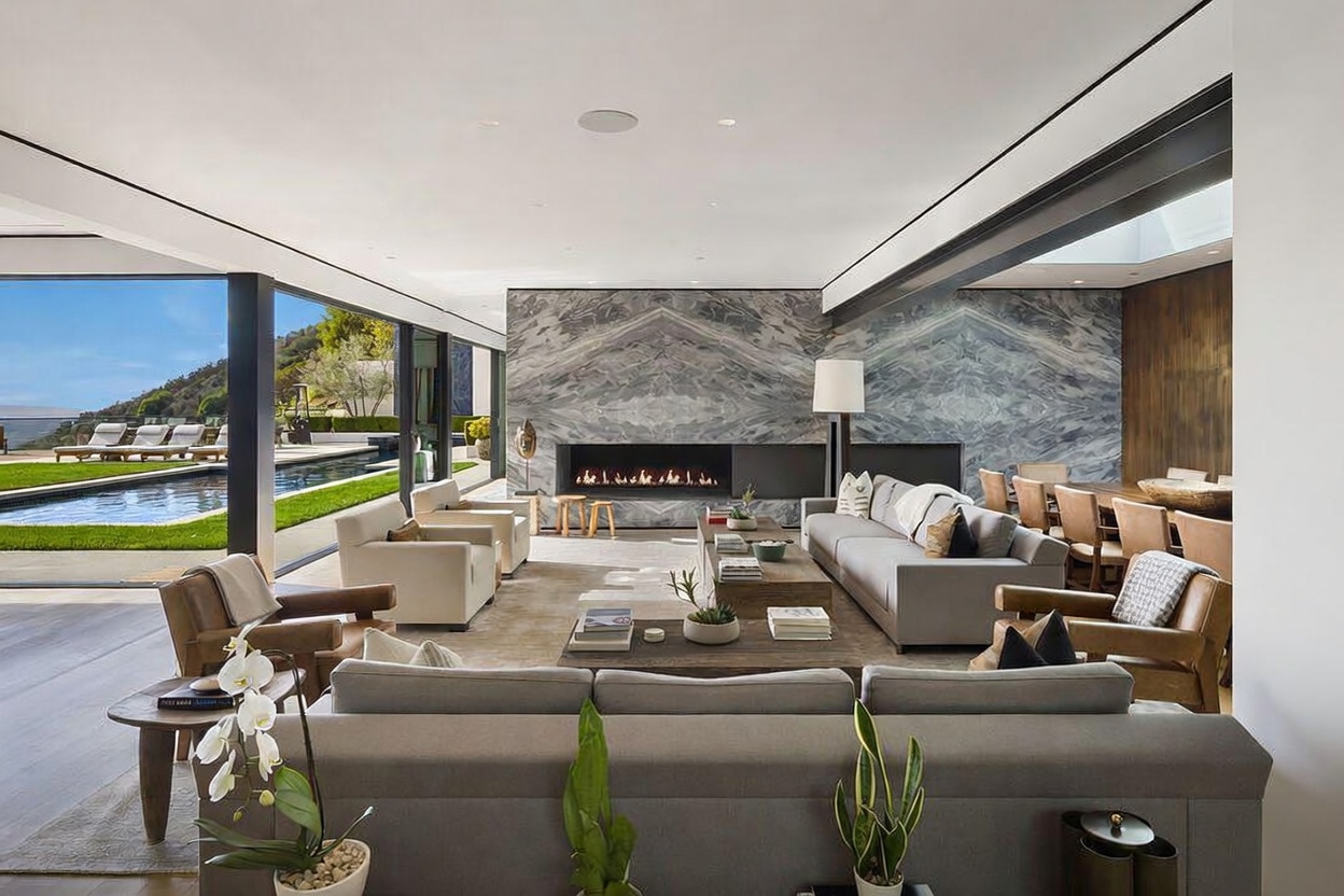 This expansive, open-concept living room features a sophisticated design with a large stone-clad chimney breast housing a linear fireplace. The space is furnished with plush grey sofas and leather armchairs, centered around a large wooden coffee table, while floor-to-ceiling glass walls provide a seamless transition to the outdoor pool area. The room is bathed in natural light, creating an airy and luxurious atmosphere that highlights the clean lines and modern aesthetic.
