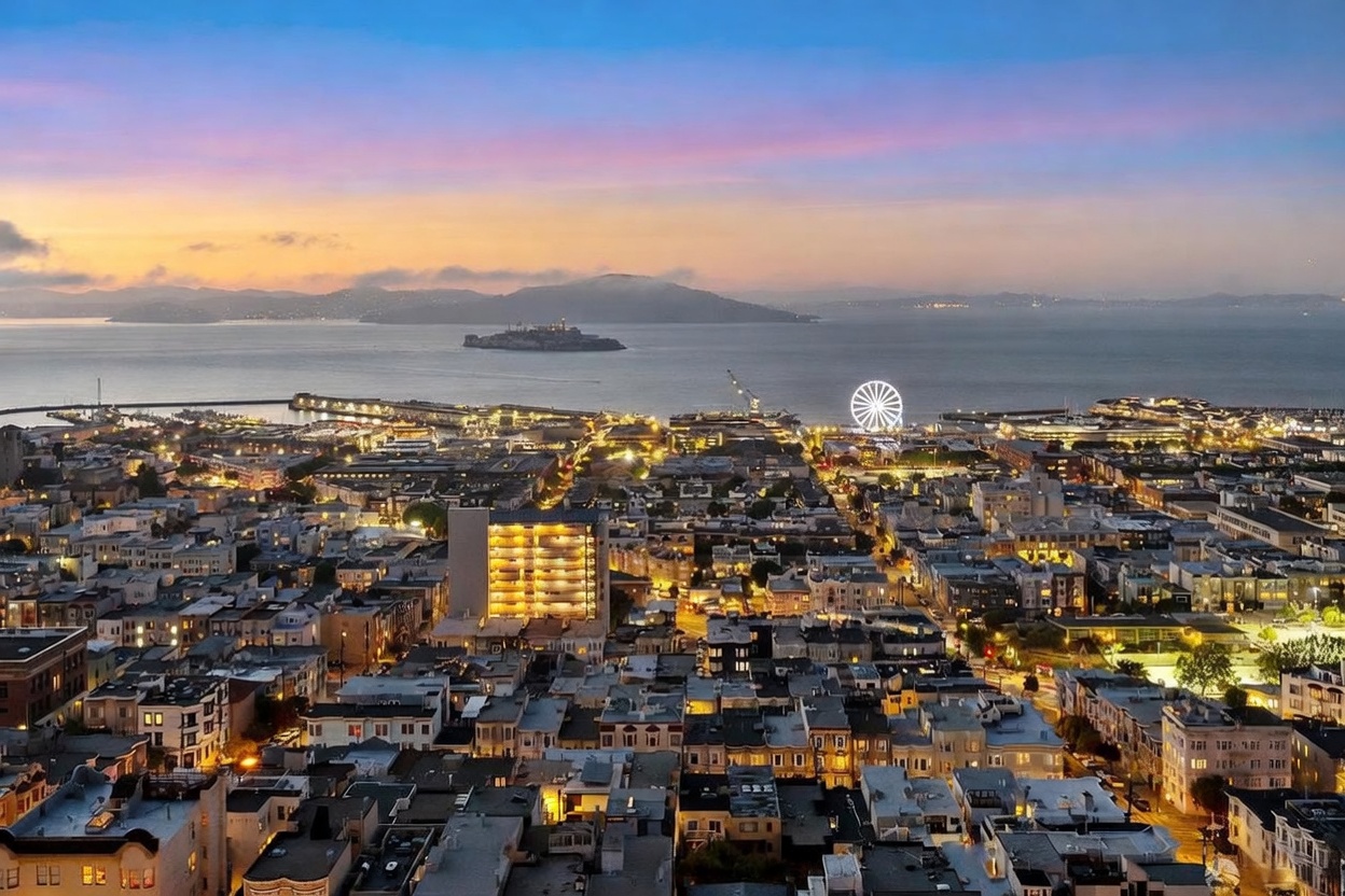 This stunning aerial view captures a sprawling cityscape at twilight, showcasing a dense urban neighborhood transitioning into the San Francisco Bay. The scene is anchored by the iconic Alcatraz Island in the distance and a brightly illuminated Ferris wheel near the waterfront, creating a vibrant and cinematic atmosphere. The warm glow of city lights contrasts beautifully with the soft, colorful hues of the sunset sky, highlighting the property's prime location and expansive views.