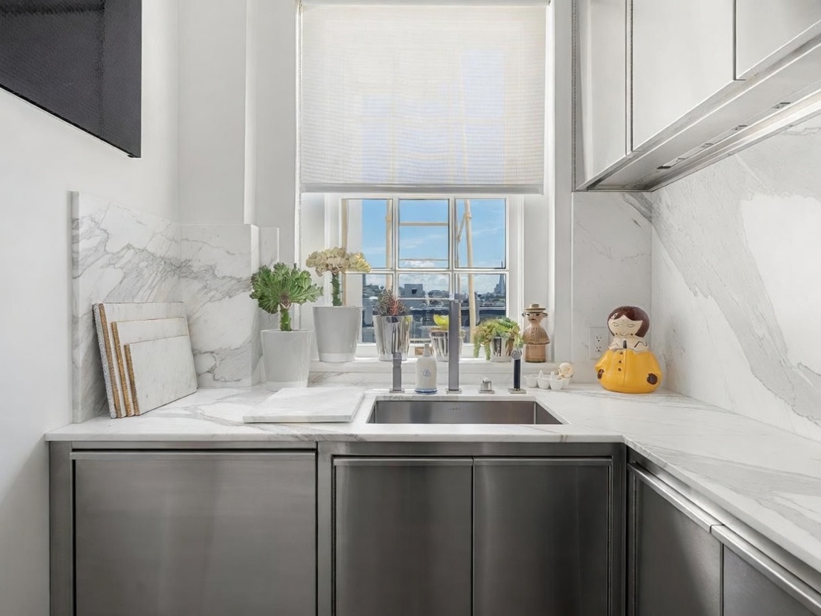 This compact, modern kitchen area features sleek stainless steel cabinetry paired with light-toned stone countertops and a matching backsplash. A central stainless steel sink sits beneath a window that provides a view of the city skyline, while a collection of decorative plants and a whimsical figurine add personality to the sill. The space is bright and clean, emphasizing a contemporary aesthetic with high-end finishes.
