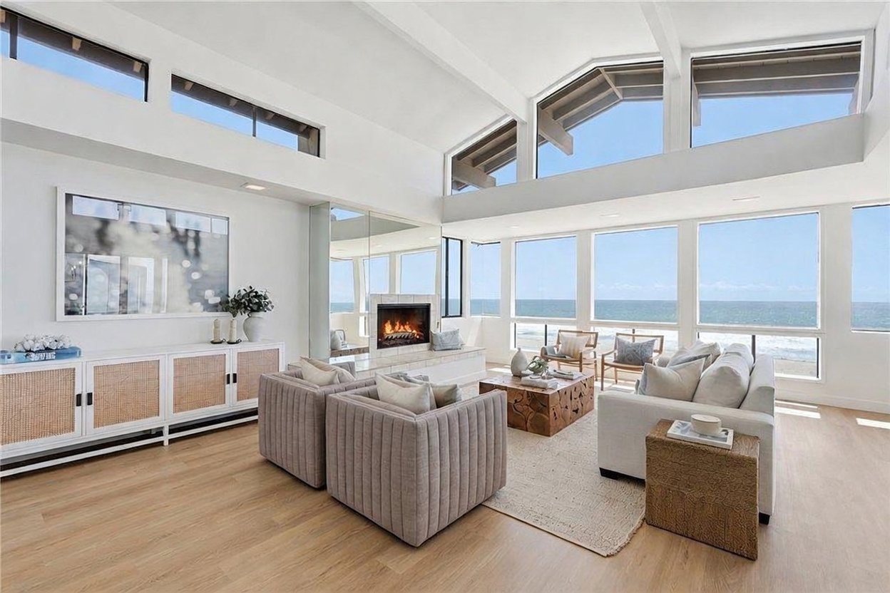 This bright and airy living room features a vaulted ceiling and expansive windows that provide a panoramic view of the ocean. The space is furnished with two textured armchairs and a large sofa arranged around a low-profile wooden coffee table, all set upon a neutral area rug. A white console with woven cabinet doors sits against the wall, complemented by a fireplace with a stone-clad chimney breast that serves as a focal point.